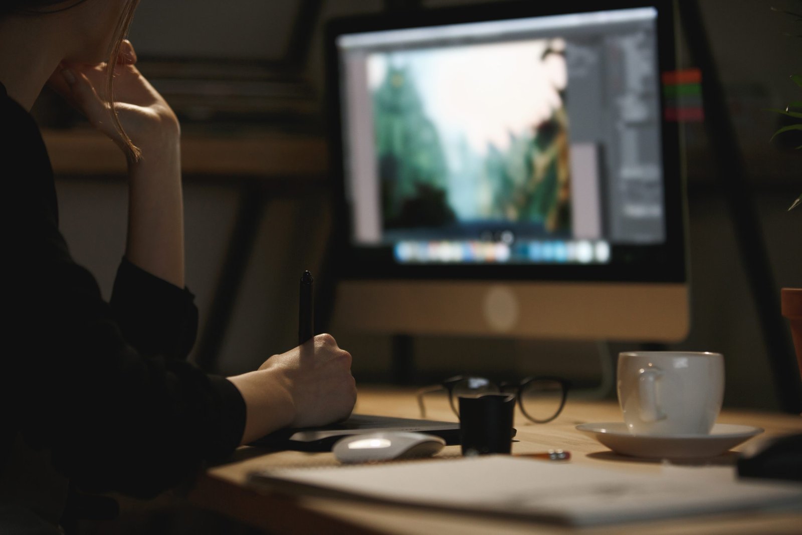 Cropped image of young lady designer sitting indoors at night using computer and graphics tablet.