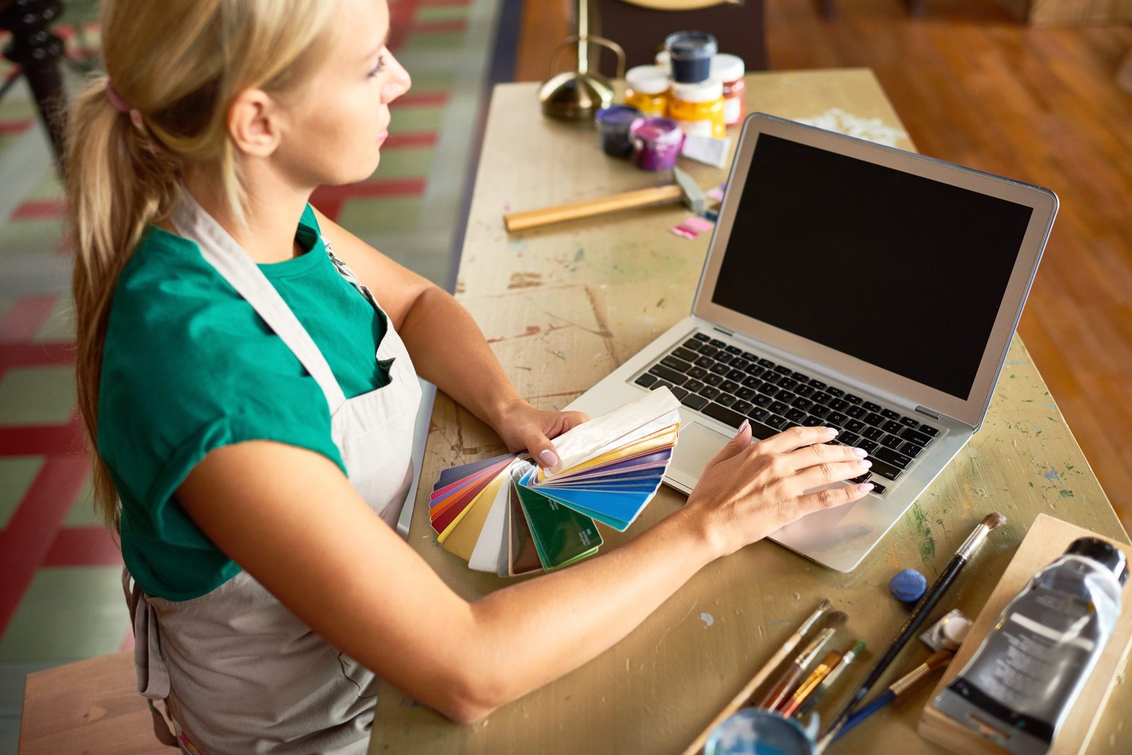 High angle portrait of pretty blond woman using laptop and choosing color palette from swatches while working in art and craft studio